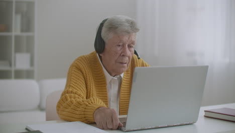 Ältere-Frau-Mit-Laptop-Computer-Ruft-Zu-Hause-Auf-Dem-Smartphone-An.-Alte-Frau-Führt-Einen-Videoanruf-Auf-Dem-Laptop,-Lächelt-Und-Redet-Glücklich-Drinnen-In-Einer-Gemütlichen-Wohnung.-Arzt-Videoanruf-älterer-Patient